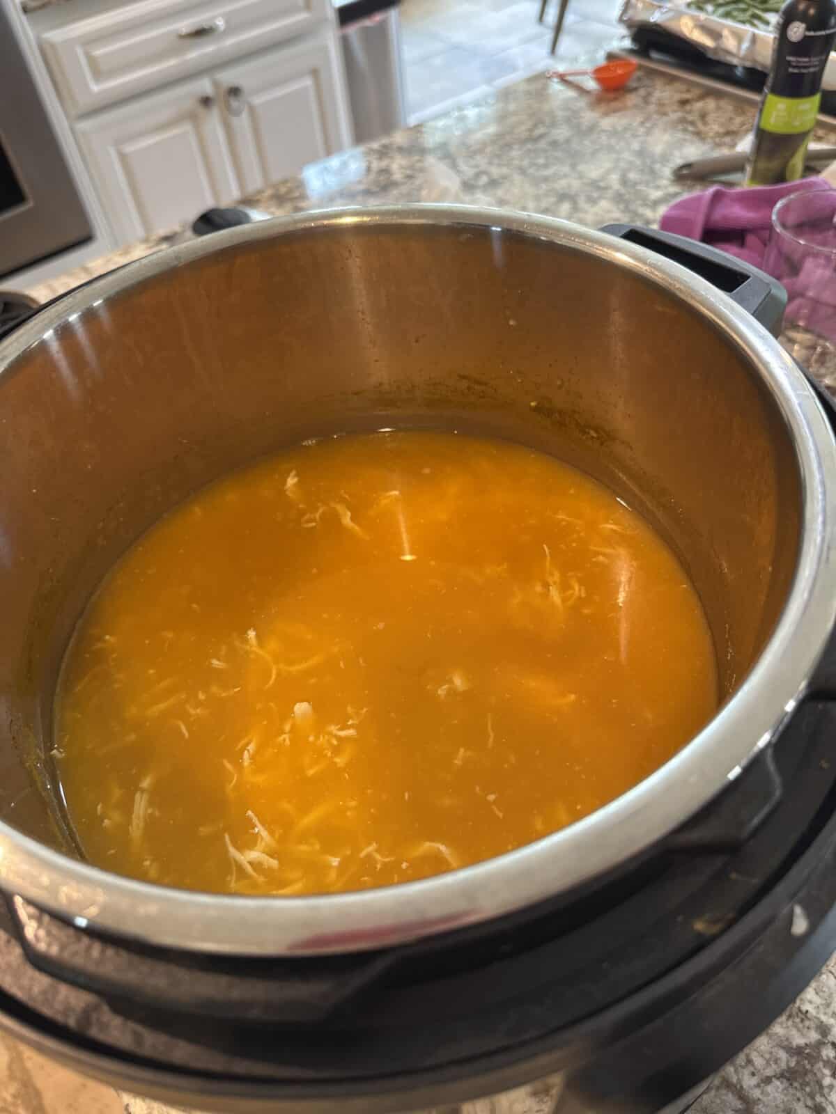 An Instant Pot filled with orange-colored Hidden Veggie Chicken Soup, featuring shredded chicken, sits on a granite kitchen counter, with cabinets and utensils visible in the background.