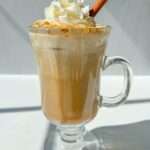 A glass mug filled with a creamy Pumpkin Spice Latte Recipe, topped with whipped cream and a cinnamon stick, sits on a white surface in bright sunlight.