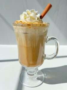 A glass mug filled with a creamy Pumpkin Spice Latte Recipe, topped with whipped cream and a cinnamon stick, sits on a white surface in bright sunlight.