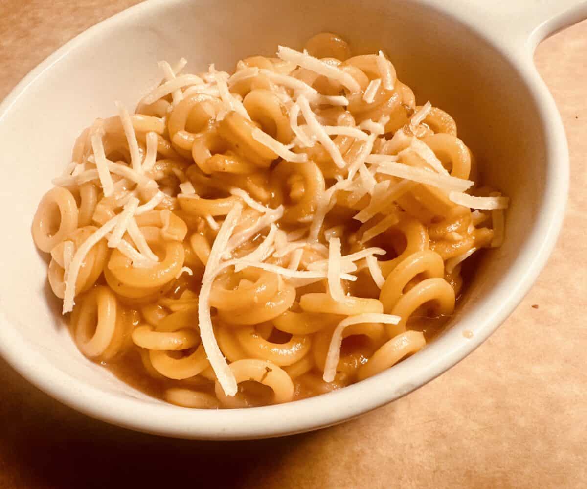 A bowl of spaghetti rings in tomato sauce topped with shredded cheese, served in a white bowl on a light brown surface.