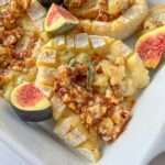 A close-up of Baked Hazelnut Brie topped with chopped nuts and honey, garnished with fresh figs and a sprig of thyme, served in a white dish.