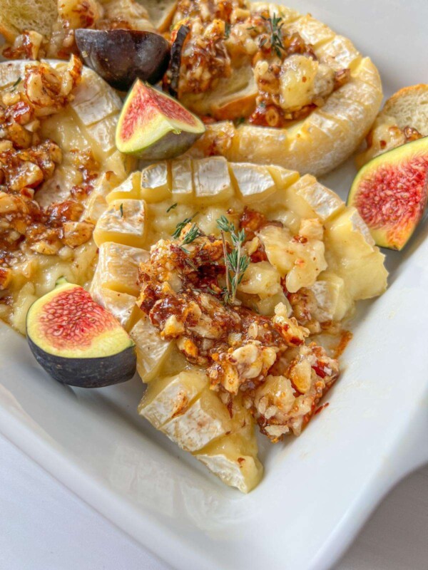 A close-up of Baked Hazelnut Brie topped with chopped nuts and honey, garnished with fresh figs and a sprig of thyme, served in a white dish.