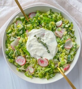 A bowl of chopped green salad with sliced radishes, topped with a dollop of creamy dressing and a sprig of thyme, is served alongside golden utensils and pairs perfectly with Bulgarian Cheese Sandwiches (Prinzesi).