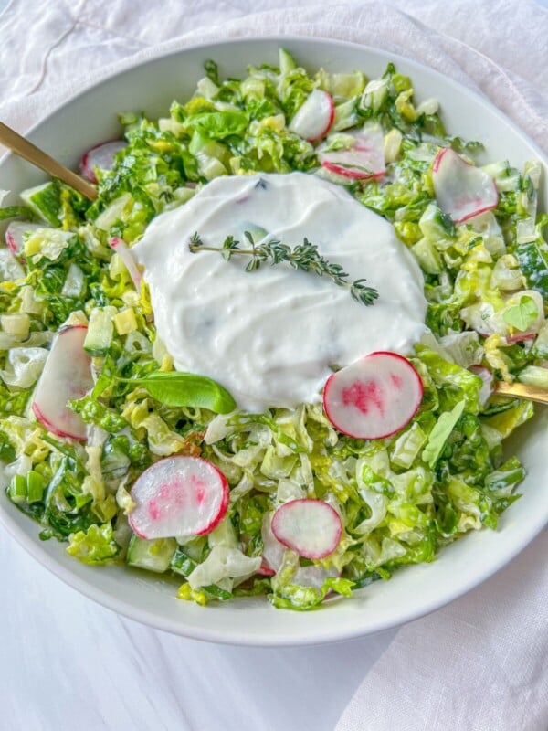A bowl of fresh chopped salad inspired by Bulgarian Salad, with lettuce, sliced radishes, and cucumbers, topped with creamy white dressing and garnished with sprigs of fresh thyme. Two gold utensils rest in the bowl.