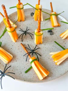 A plate of Cheese Broomsticks—cheese sticks arranged as broomsticks with pretzel handles and green ties—adorned with plastic toy spiders, on a speckled plate.