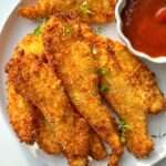 A plate of crispy chicken fingers, golden-brown and breaded, garnished with fresh thyme and served with a side of vibrant red dipping sauce in a small white bowl.