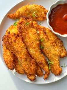 A plate of crispy chicken fingers, golden-brown and breaded, garnished with fresh thyme and served with a side of vibrant red dipping sauce in a small white bowl.