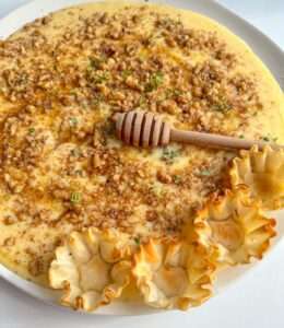 A plate of creamy Custard Baklava dessert topped with chopped nuts and honey, garnished with a honey dipper and served with three crispy pastry cups on the side.
