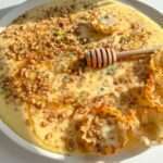 A plate of creamy custard baklava dessert topped with chopped nuts, a drizzle of honey, and small crispy pastry cups. A wooden honey dipper rests on top, and fresh herbs are sprinkled as garnish.