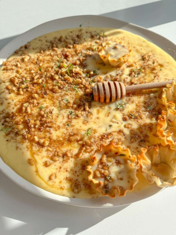 A plate of creamy custard baklava dessert topped with chopped nuts, a drizzle of honey, and small crispy pastry cups. A wooden honey dipper rests on top, and fresh herbs are sprinkled as garnish.