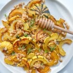 Roasted Delicata Squash slices on a white plate, topped with chopped nuts, fresh herbs, and a honey dipper drizzling golden honey over this vibrant roasted delicata squash dish.