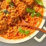 A white pan filled with homemade Spaghettios in red tomato sauce, topped with fresh basil leaves. A wooden spoon rests in the pasta, while sunlight casts gentle shadows across the inviting dish.