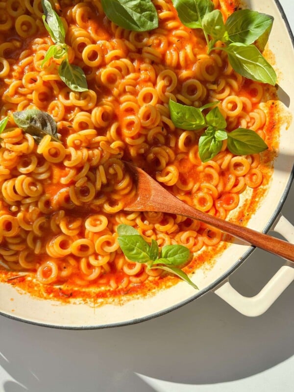 A white pan filled with homemade Spaghettios in red tomato sauce, topped with fresh basil leaves. A wooden spoon rests in the pasta, while sunlight casts gentle shadows across the inviting dish.