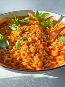 A bowl of homemade Spaghettios in tomato sauce, garnished with fresh basil leaves, with a wooden spoon resting inside. Sunlight casts soft shadows on the white surface below.