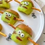 Sliced kiwis decorated as Frankenstein faces with candy eyes, chocolate-drawn mouths and scars, kiwi skin as hair, and pretzel sticks as bolts are a spooky treat—perfect alongside halloween mummy hotdogs for your festive celebration.