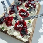 A Halloween Cheese Board features a wooden board topped with whipped cream cheese, red jam, candy eyes, and spooky plastic spiders and skeletons. Silver spreader knives complete this eerie, Halloween-themed appetizer.