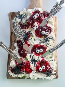 A Halloween Cheese Board topped with whipped cream, red jelly, candy eyes, and black gummy spiders, with three spreader knives featuring skeleton handles arranged on top for a spooky Halloween-themed treat.
