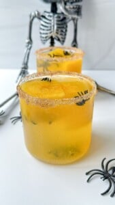 A yellow cocktail with an orange garnish, sugar on the rim, and small fake spiders sits in front of a decorative skeleton, while a Strawberry Mojito and more spiders add to the festive scene in the background.