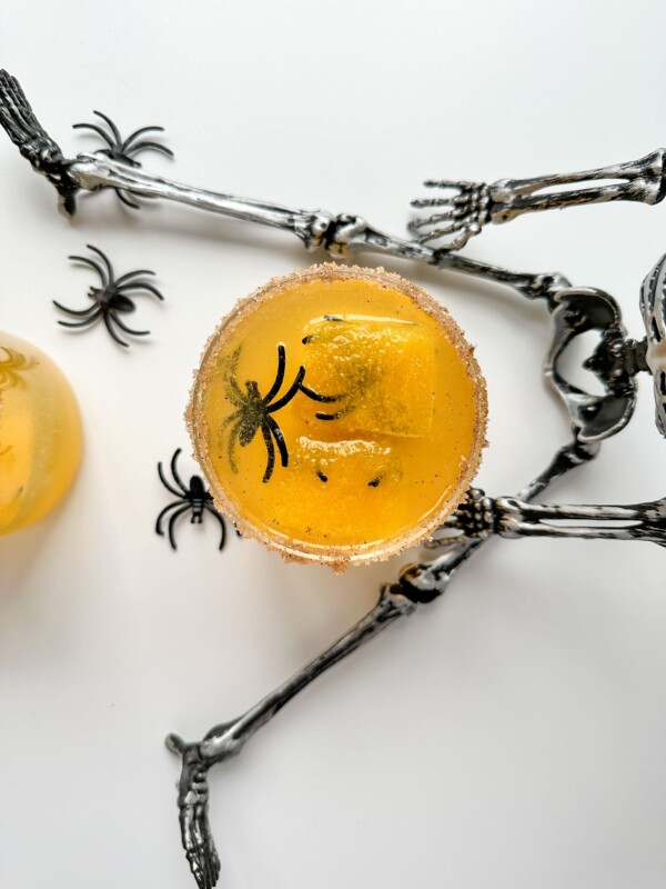 A top view of a yellow drink with a sugared rim and plastic black spider decorations, placed on a silver skeleton prop, showcases one of the best simple Halloween cocktails in a festive, spooky setting.