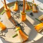A plate features Cheese Broomsticks—snacks made of string cheese, pretzel sticks, and green herbs—decorated with plastic spiders for a festive Halloween touch.