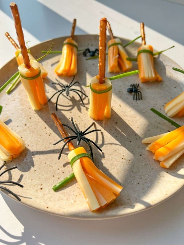 A plate features Cheese Broomsticks—snacks made of string cheese, pretzel sticks, and green herbs—decorated with plastic spiders for a festive Halloween touch.