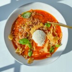 A white plate of Lasagna Soup with wavy noodles, ground meat, red tomato broth, fresh basil, and a scoop of ricotta cheese is served with a gold spoon on a sunlit white table.
