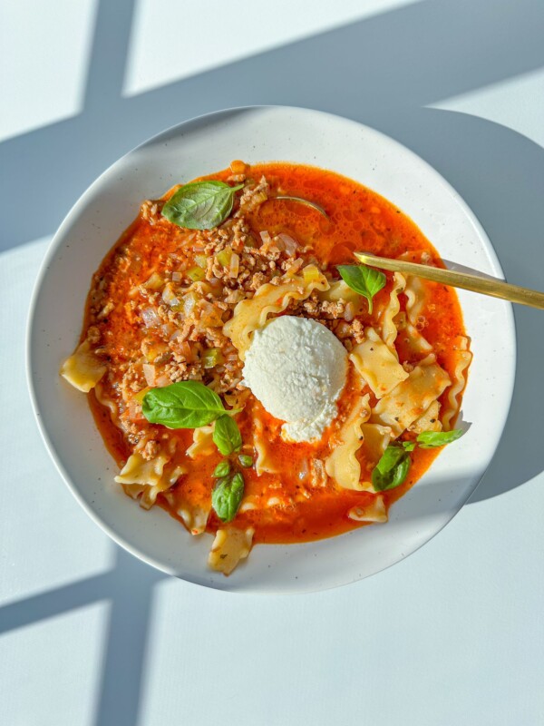 A white plate of Lasagna Soup with wavy noodles, ground meat, red tomato broth, fresh basil, and a scoop of ricotta cheese is served with a gold spoon on a sunlit white table.