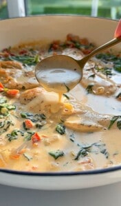 A spoon scoops creamy soup from a bowl, filled with chunks of vegetables and greens. The soup's surface shows a rich, smooth texture with hints of herbs, offering a warm, inviting appearance.
