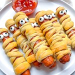 A plate of halloween mummy hotdogs wrapped in crescent dough with candy eyes on top. A small bowl of ketchup sits behind them, perfect for dipping.