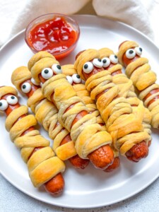 A plate of halloween mummy hotdogs wrapped in crescent dough with candy eyes on top. A small bowl of ketchup sits behind them, perfect for dipping.