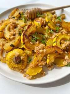 A plate of Roasted Delicata Squash and yellow pattypan squash, topped with chopped walnuts, fresh herbs, and a drizzle of honey. A honey dipper rests on the plate.