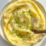 A plate of creamy Roasted Garlic Mashed Potatoes topped with butter, cracked black pepper, and fresh green herbs, with a wooden spoon resting in the dish.