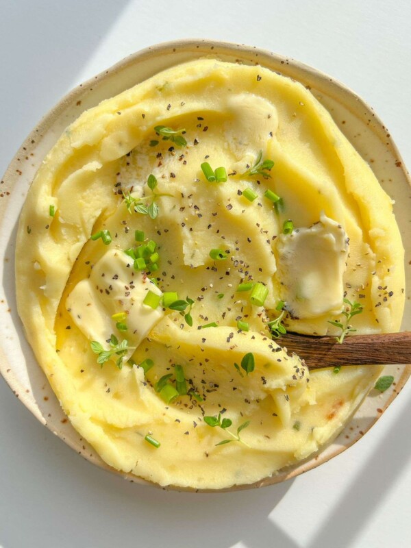 A plate of creamy Roasted Garlic Mashed Potatoes topped with butter, cracked black pepper, and fresh green herbs, with a wooden spoon resting in the dish.