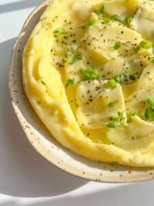 A bowl of creamy roasted garlic mashed potatoes topped with butter, chopped chives, fresh herbs, and cracked black pepper, served in a speckled ceramic dish under bright natural light.