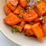 A speckled ceramic plate filled with Roasted Sweet Potatoes, garnished with fresh green herbs. The sweet potatoes are golden, slightly caramelized, and bursting with savory-sweet flavor.