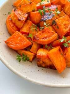A speckled ceramic plate filled with Roasted Sweet Potatoes, garnished with fresh green herbs. The sweet potatoes are golden, slightly caramelized, and bursting with savory-sweet flavor.