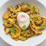 A white plate features a Delicata Squash Salad with roasted delicata squash slices, sautéed onions, fresh basil leaves, and a poached egg in the center, all artfully arranged on a white background.