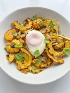 A white plate features a Delicata Squash Salad with roasted delicata squash slices, sautéed onions, fresh basil leaves, and a poached egg in the center, all artfully arranged on a white background.