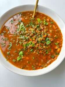 A bowl of hearty lentil soup, known as The Best Lentil Soup, with chopped vegetables and fresh herbs, garnished with basil leaves and a spoon resting in the bowl.