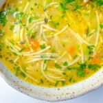 A close-up of a bowl of clear Turkey Noodle Soup with thin noodles, diced carrots, fresh herbs, and a light broth, served in a speckled ceramic bowl.