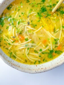 A close-up of a bowl of clear Turkey Noodle Soup with thin noodles, diced carrots, fresh herbs, and a light broth, served in a speckled ceramic bowl.