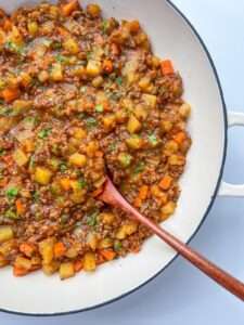 A close-up of a white pan filled with a hearty ground beef stew, reminiscent of Moussaka with Potatoes, featuring diced potatoes, carrots, and herbs, with a wooden spoon resting inside the mixture.