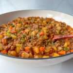 A close-up of a bowl filled with a chunky stew resembling Moussaka with Potatoes, featuring ground meat, diced potatoes, carrots, and green herbs, with a wooden spoon resting inside. The dish appears hearty and freshly prepared.