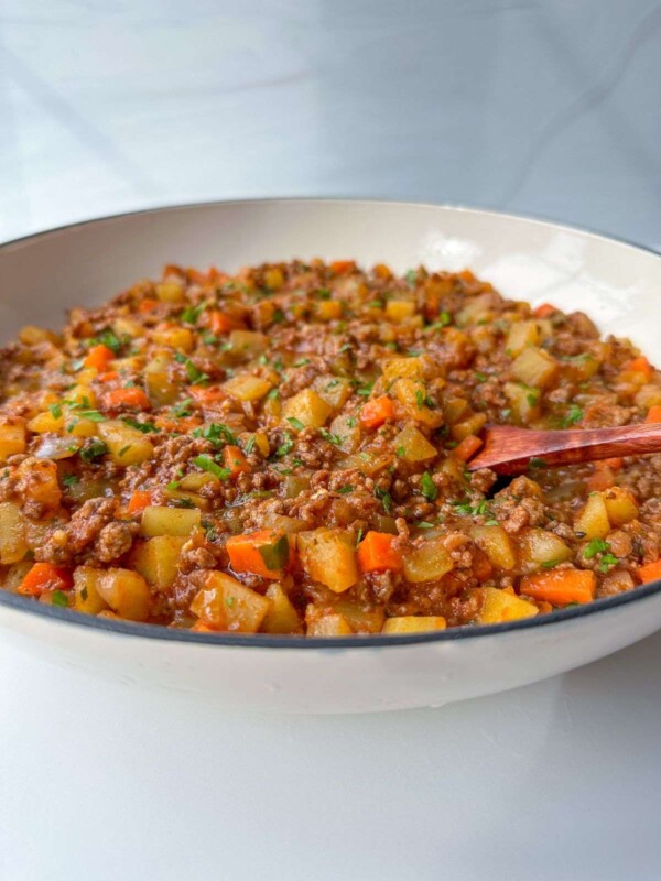 A close-up of a bowl filled with a chunky stew resembling Moussaka with Potatoes, featuring ground meat, diced potatoes, carrots, and green herbs, with a wooden spoon resting inside. The dish appears hearty and freshly prepared.
