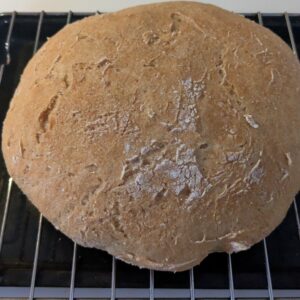 A round loaf of rustic, homemade bread with a golden-brown crust rests on a wire rack over a black baking tray. The surface is textured with cracks and a light dusting of flour.