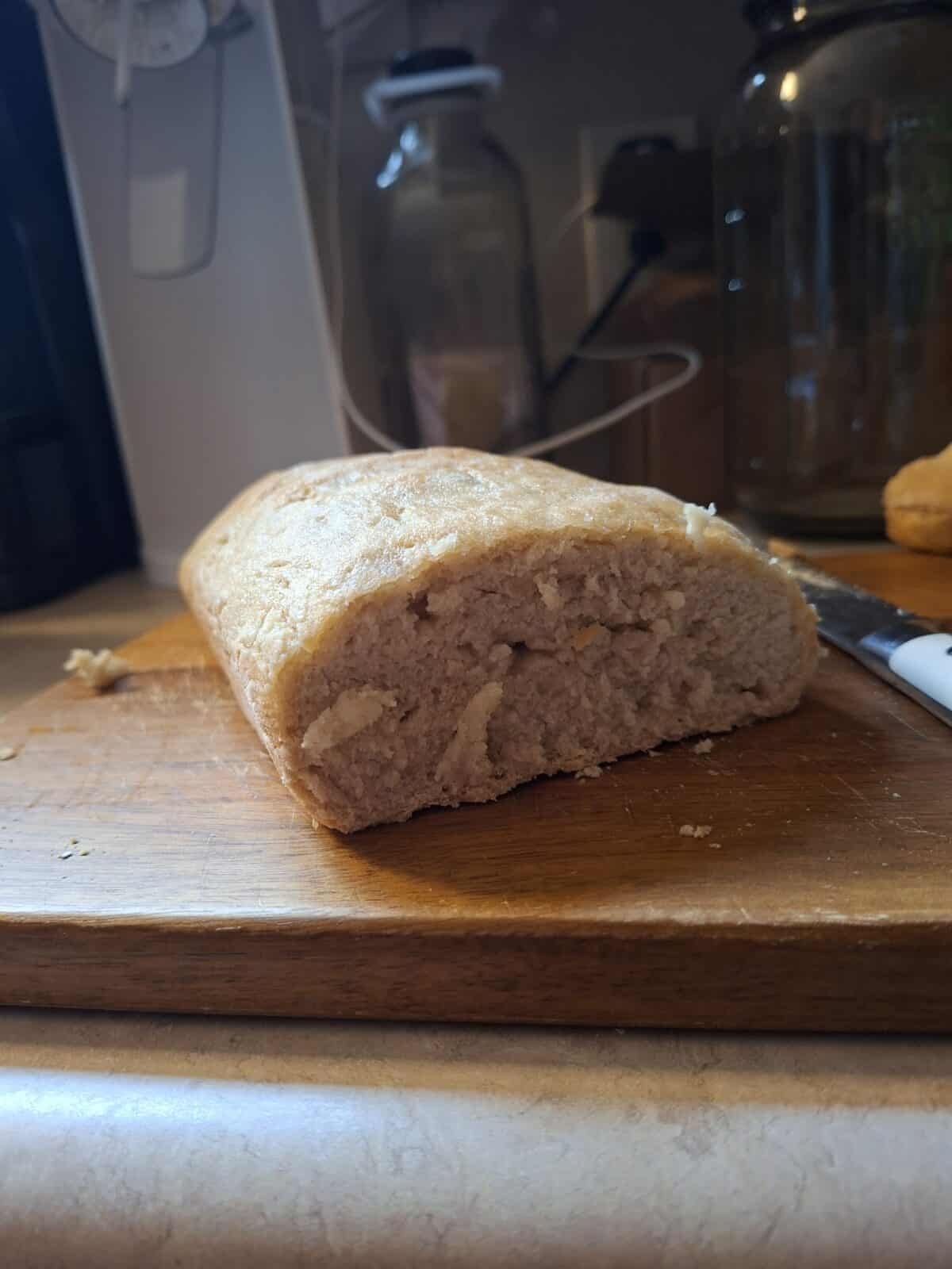 A loaf of No Knead Gluten Free Bread with a dense, uneven texture sits partially sliced on a wooden cutting board, with a bread knife and glass jars visible in the background on a kitchen counter.