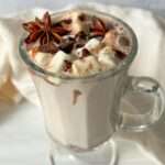 A glass mug filled with creamy hot chocolate made from Kinder Hot Chocolate Bombs, topped with marshmallows, chocolate shavings, and star anise, sits on a white surface with a light-colored cloth in the background.