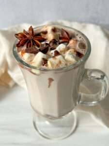 A glass mug filled with creamy hot chocolate made from Kinder Hot Chocolate Bombs, topped with marshmallows, chocolate shavings, and star anise, sits on a white surface with a light-colored cloth in the background.