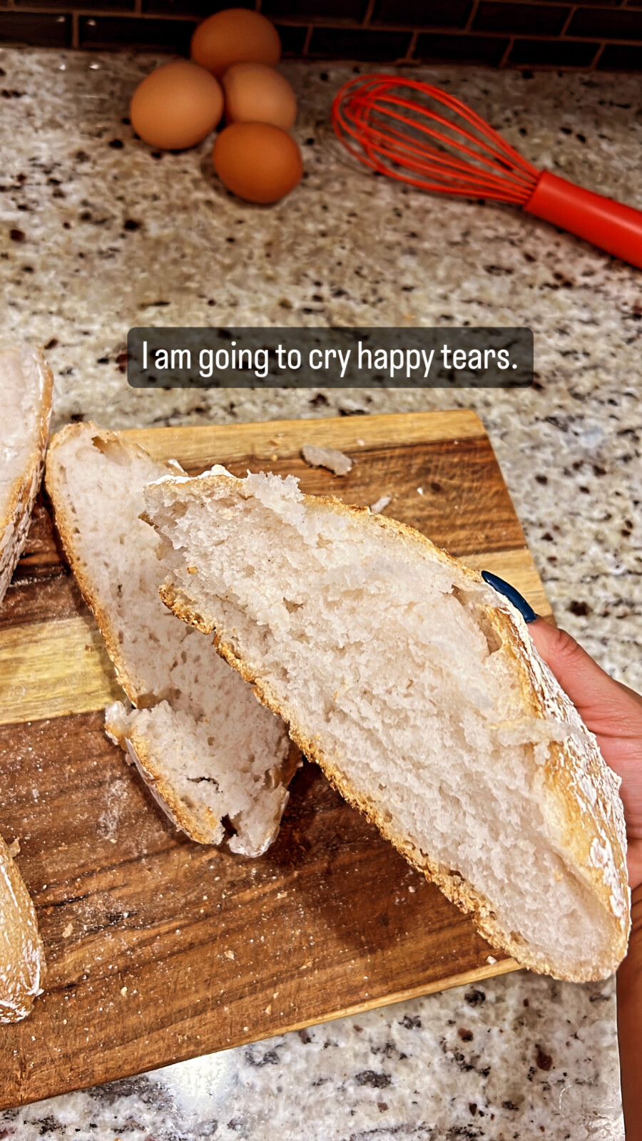 A hand holds a slice of rustic No Knead Gluten Free Bread over a wooden cutting board, with more bread slices, brown eggs, and a red whisk on a speckled countertop. Text overlay reads, "I am going to cry happy tears.