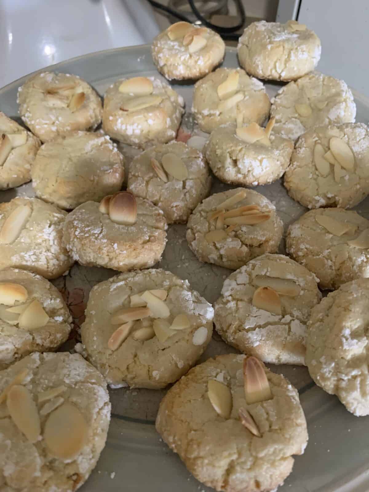 A plate of round, crinkle-textured Italian Amaretti Cookies topped with sliced almonds, arranged closely together. The cookies have a light golden color and a dusting of powdered sugar.